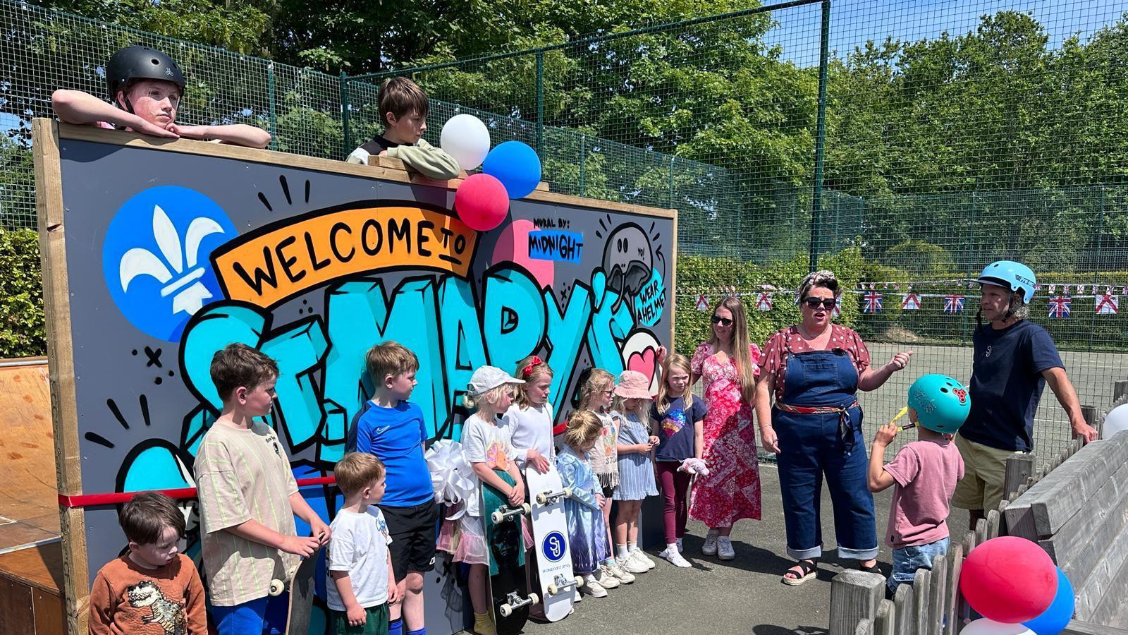 A group of children and adults are gathered outdoors in front of a colorful graffiti-style sign that reads 'Welcome to St. Mary's.' The sign features a blue fleur-de-lis symbol and additional text saying 'Made by Moonlight' and 'Skate Park.' Some children are holding skateboards, and balloons are attached to the sign. It’s a sunny day with trees and a fence in the background.