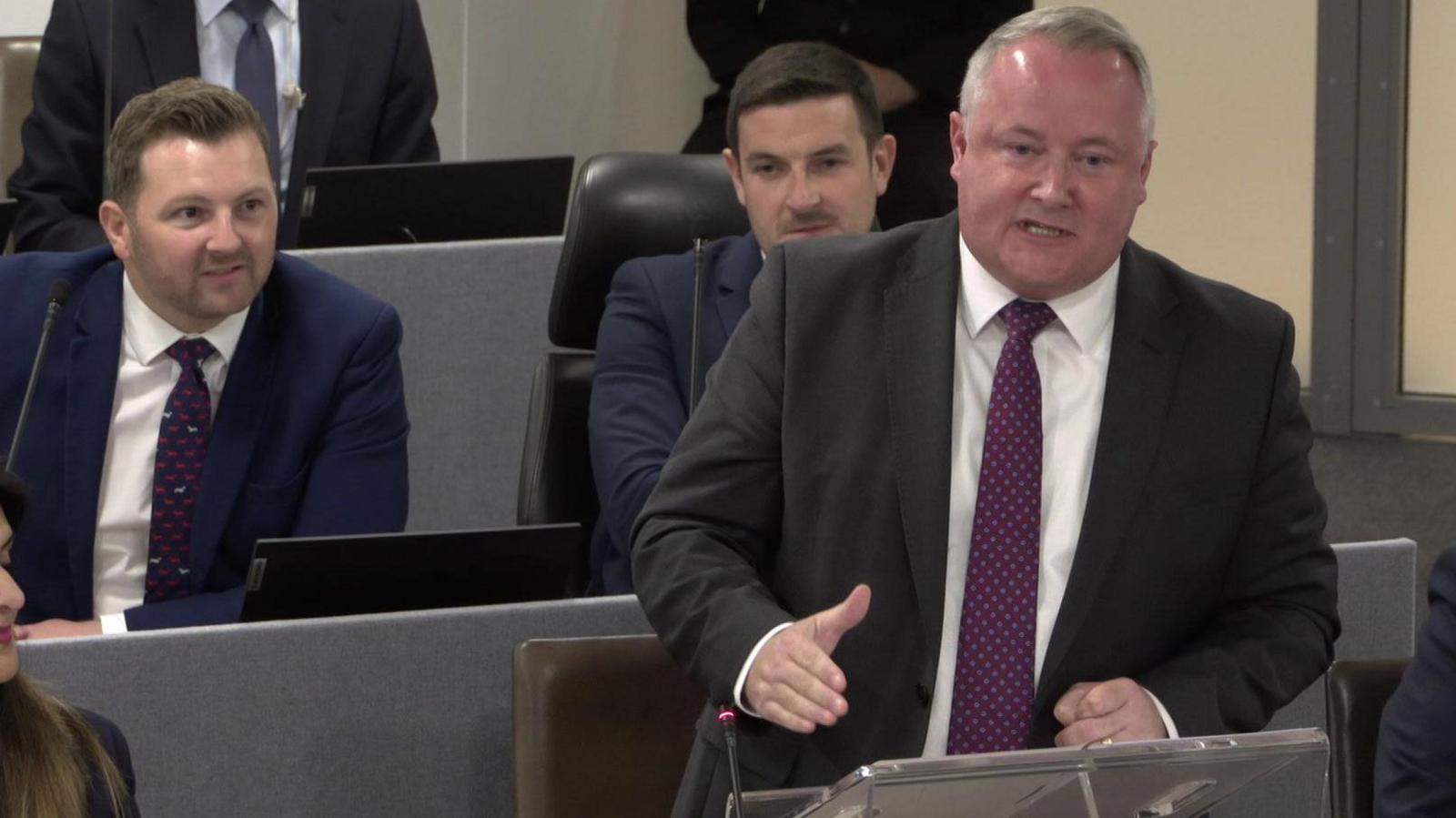 Darren Millar on his feet in the Senedd addressing Eluned Morgan during First Minister's Questions. Fellow Conservative members are seated behind him and either side, some partially out of shot.