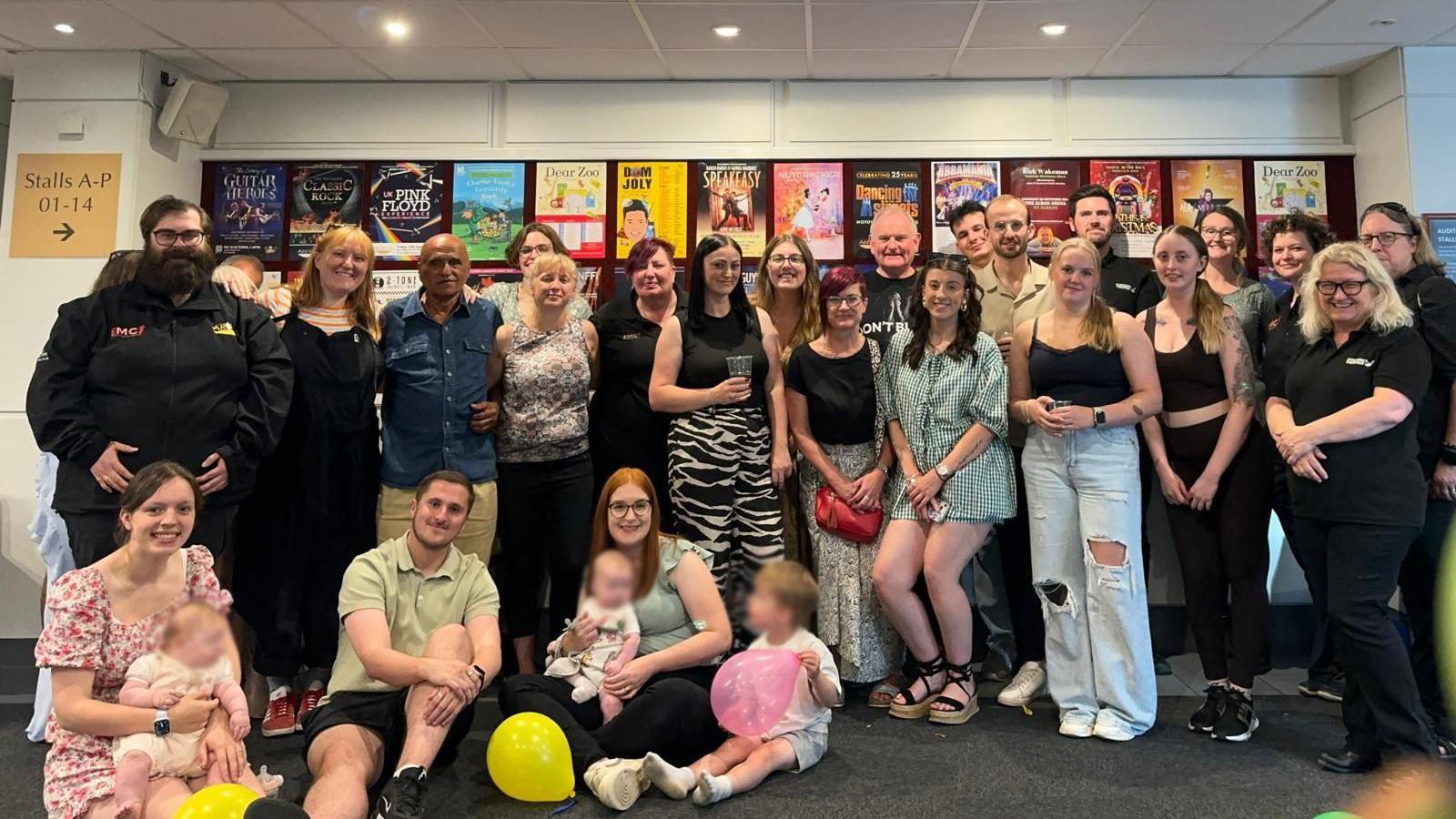 A large group of people gathering to celebrate 50 years of service of Mick Chaudry. It shows several people standing up, three people sitting down, with young children on their lap, looking at balloons.