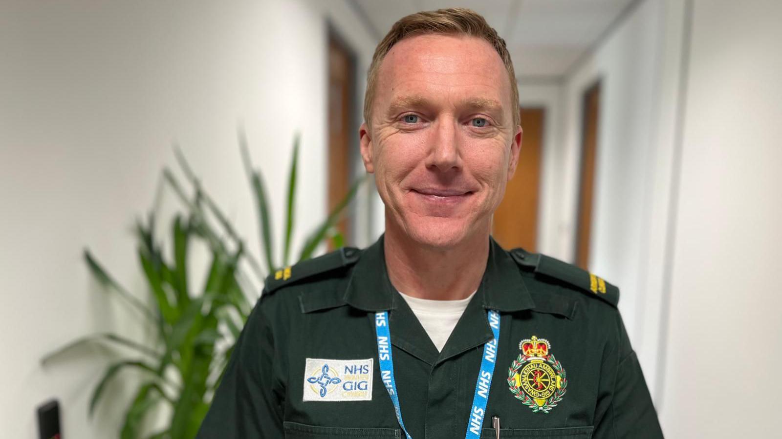 Ed is wearing a green ambulance service uniform and is stood in an office corridor with a plant in the background.