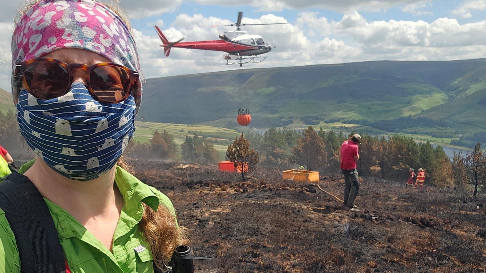 A selfie by a woman in protective clothing in front of a helicopter flying in a load of water to a burnt landscape