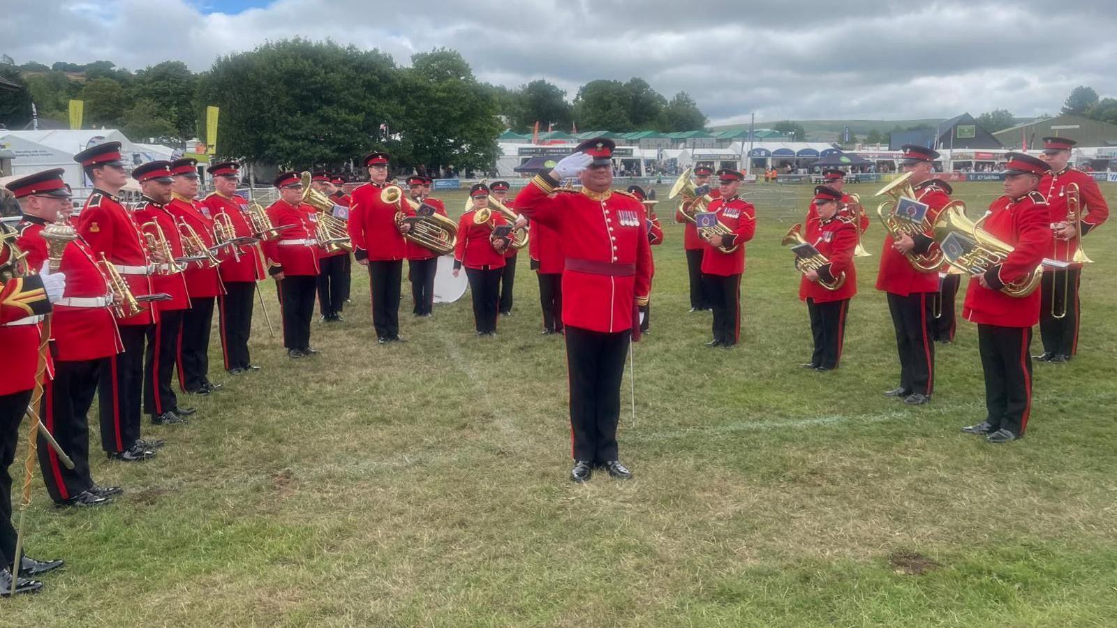 A wide image of the Regimental Band performing on a field. There is around 30 of them and they are all wearing their red uniform with their medals on display. All the band members are holding their brass musical instruments. 