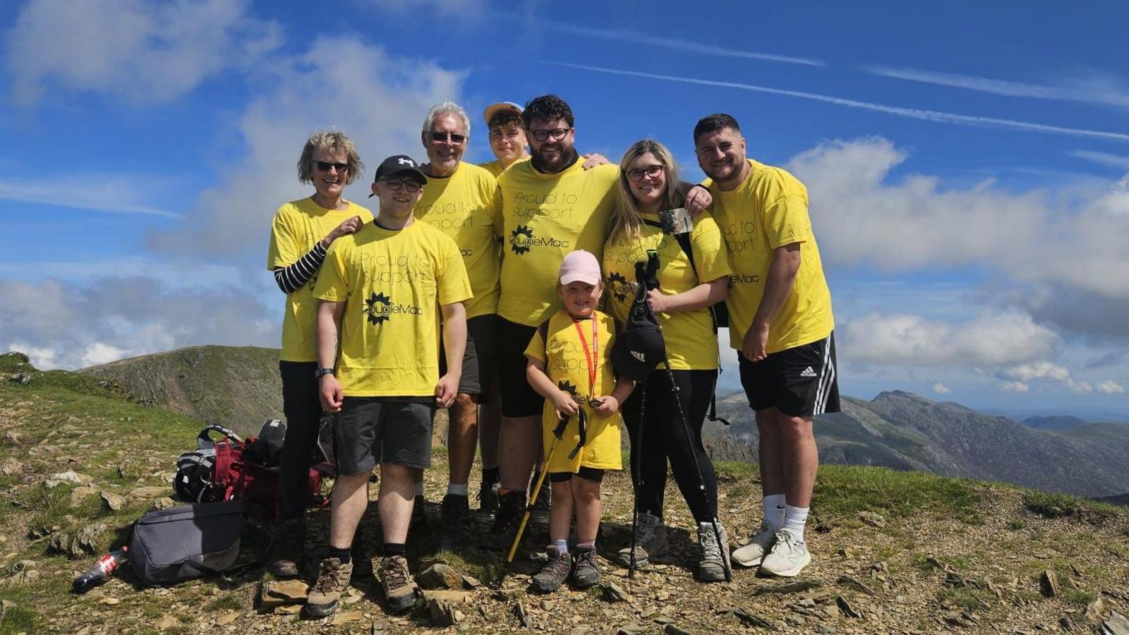 A group of people wearing yellow t-shirts at the top of a mountain, posing for the photograph. There are rucksacks and some ae carrying hiking poles.