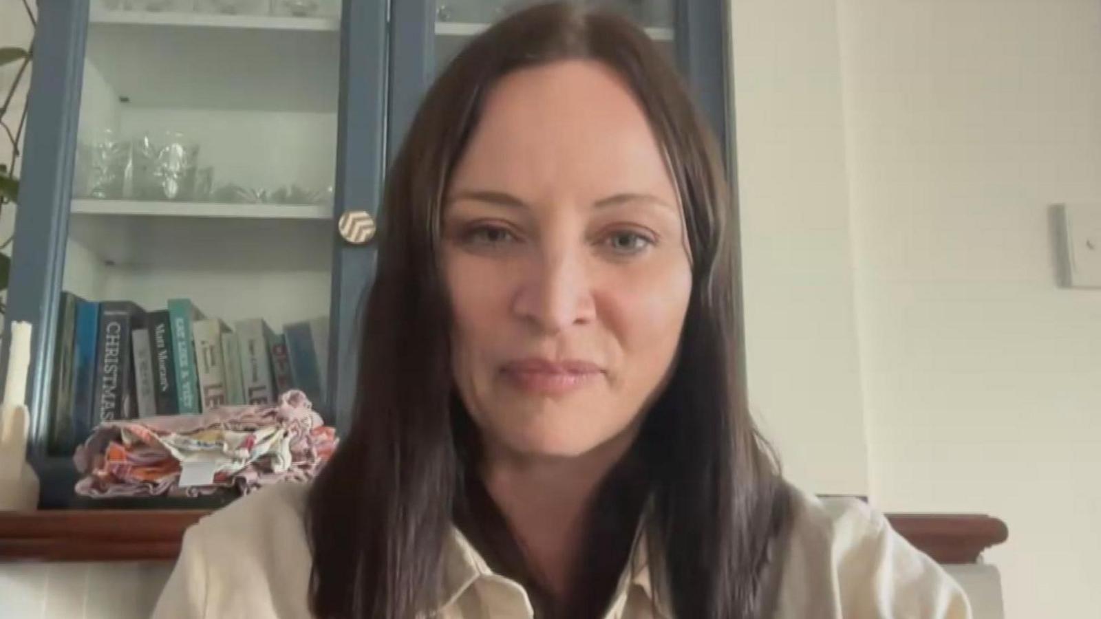 Alex sits on a video call. She is wearing a white shirt and has shoulder-length black her. Behind her is a mantlepiece with books and glasses on show.