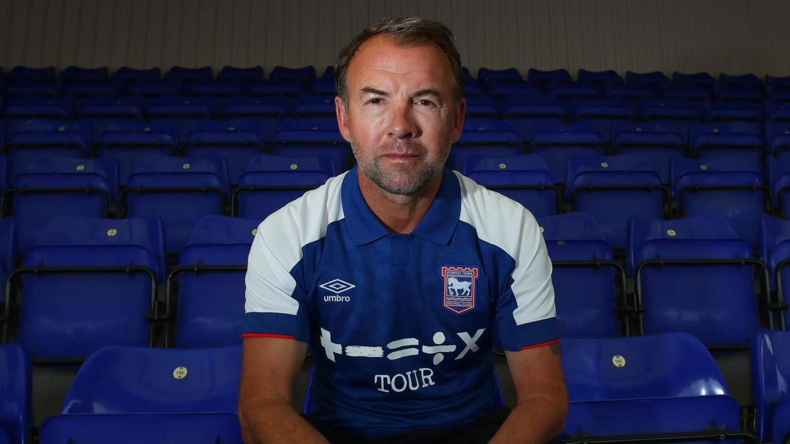 Marcus Stewart sits in an empty football stadium on a blue seat and is pictured in a blue and white Ipswich Town shirt.