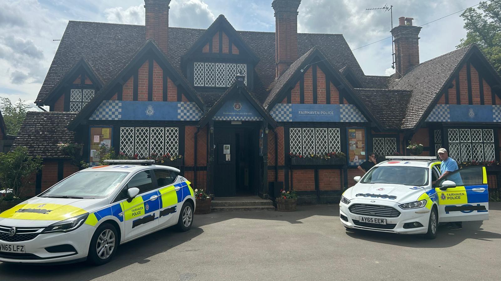 What looks like a police station with brick walls, white lattice windows, and multiple chimneys, with two police cars parked in front