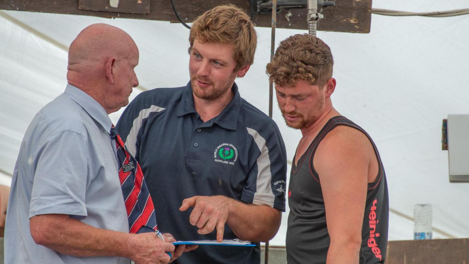 Aaron Magee is standing in the middle wearing a navy t-shirt. He is pointing at a clip board which has a sheet of paper on it. He is looking at a man with a bald head wearing a light blue shirt and red, white and blue tie. There is also a man in a black vest top looking at the clip board. The background is white. 