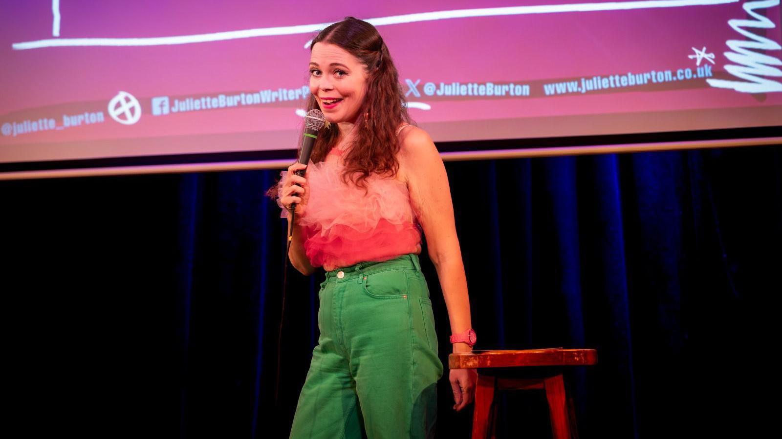 Juliette Burton on stage with a microphone doing stand-up comedy, wearing bright green jeans and a pink ruffled top.  There is a red stool next to her.