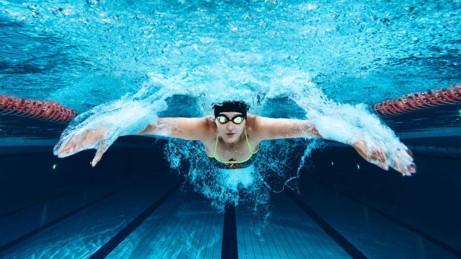 A woman is swimming underwater in a pool - she's wearing a black swimming cap and googles