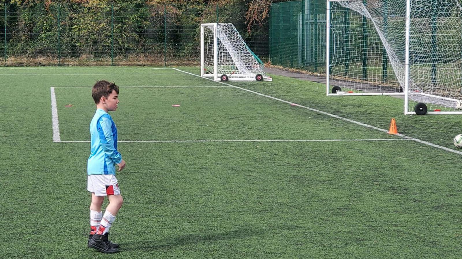 Reuben is standing on a synthetic turf football pitch, dressed in a blue long-sleeve top, white shorts with red trim, and black boots with white socks. Behind him, two small goals are set up, one with its net partially collapsed. Orange training cones are scattered across the field.
