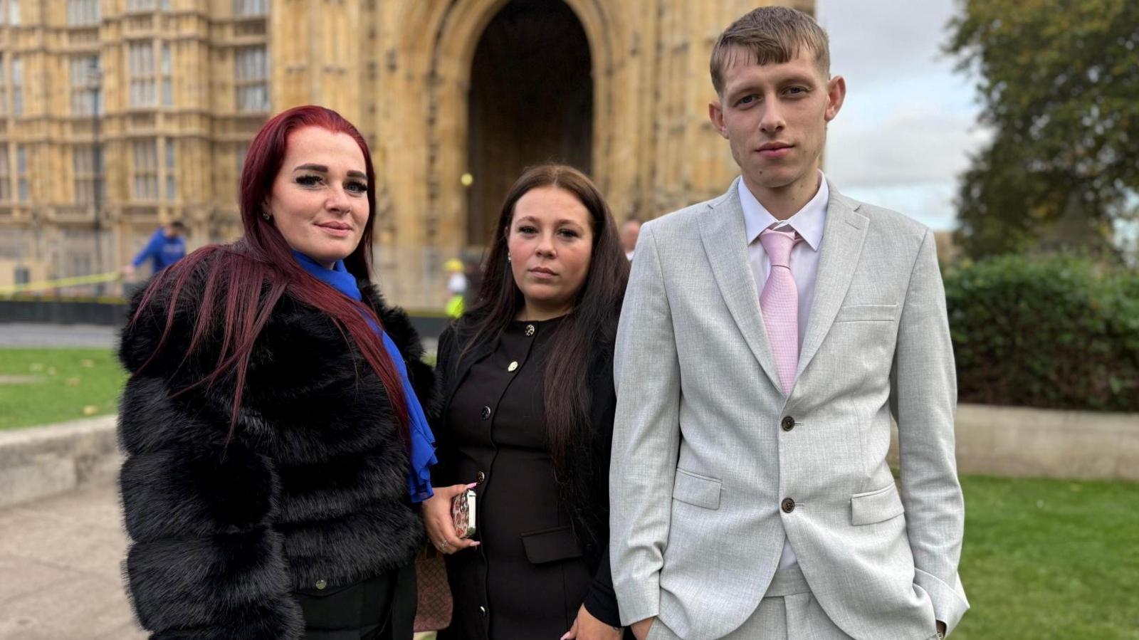 Two women and a man stood together on grass outside. The woman on the left is wearing a black outfit with a fur-style coat and a blue scarf. She has long, dark hair.  The woman in the middle is dressed in a black dress with gold buttons and is holding a phone in one hand. The man on the right is wearing a light grey suit with a pink tie and white shirt. He has one hand in the pocket of the trousers.