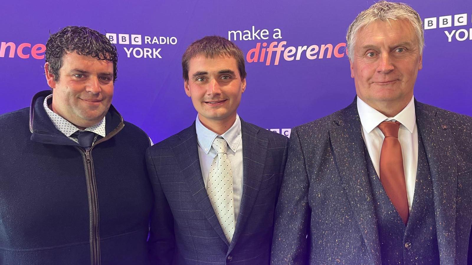 Three men standing in front of a purple background marked BBC Radio York, Make A Difference