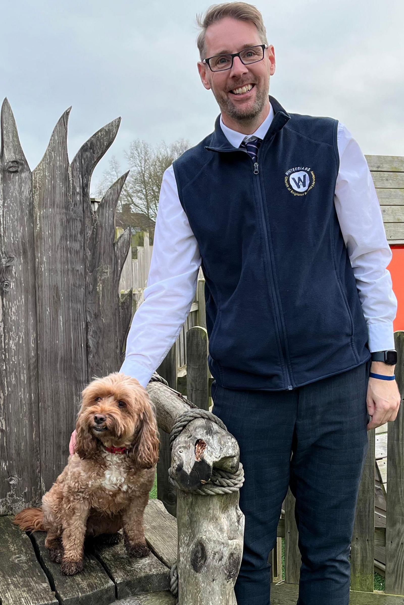 Head teacher Mat Tuckwood stands next to Poppy the cockapoo. He is wearing navy trousers and a navy fleece with a light blue shirt under it. He wears glasses and is very tall. 