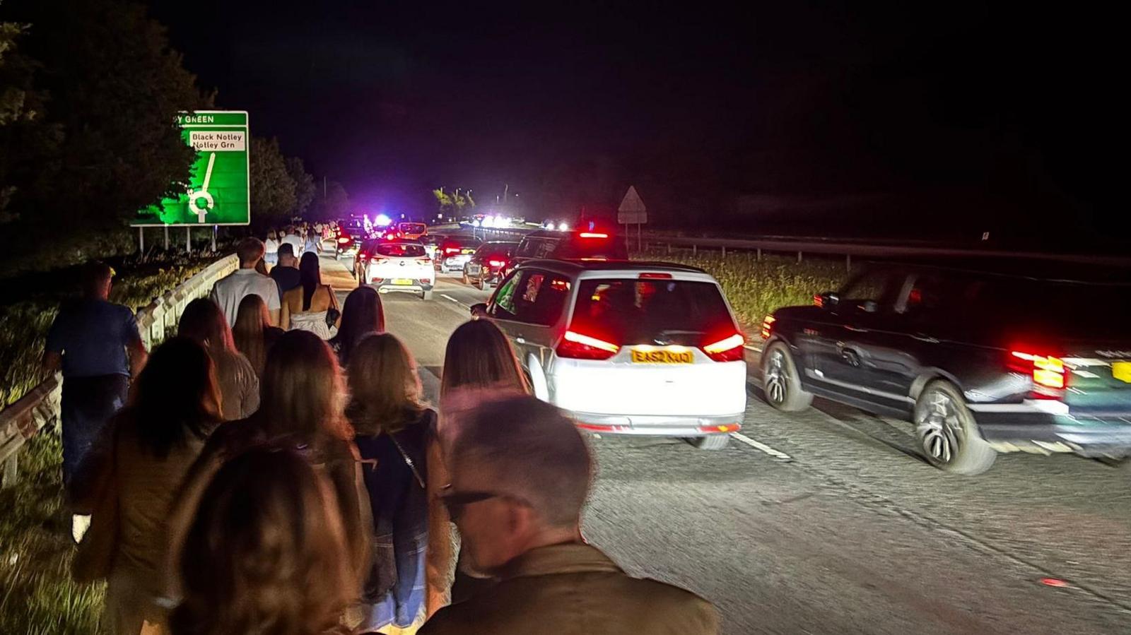 People are walking on a pavement, which is on the left. On the right, there is a busy road with traffic. It is dark. The cars have their lights on.