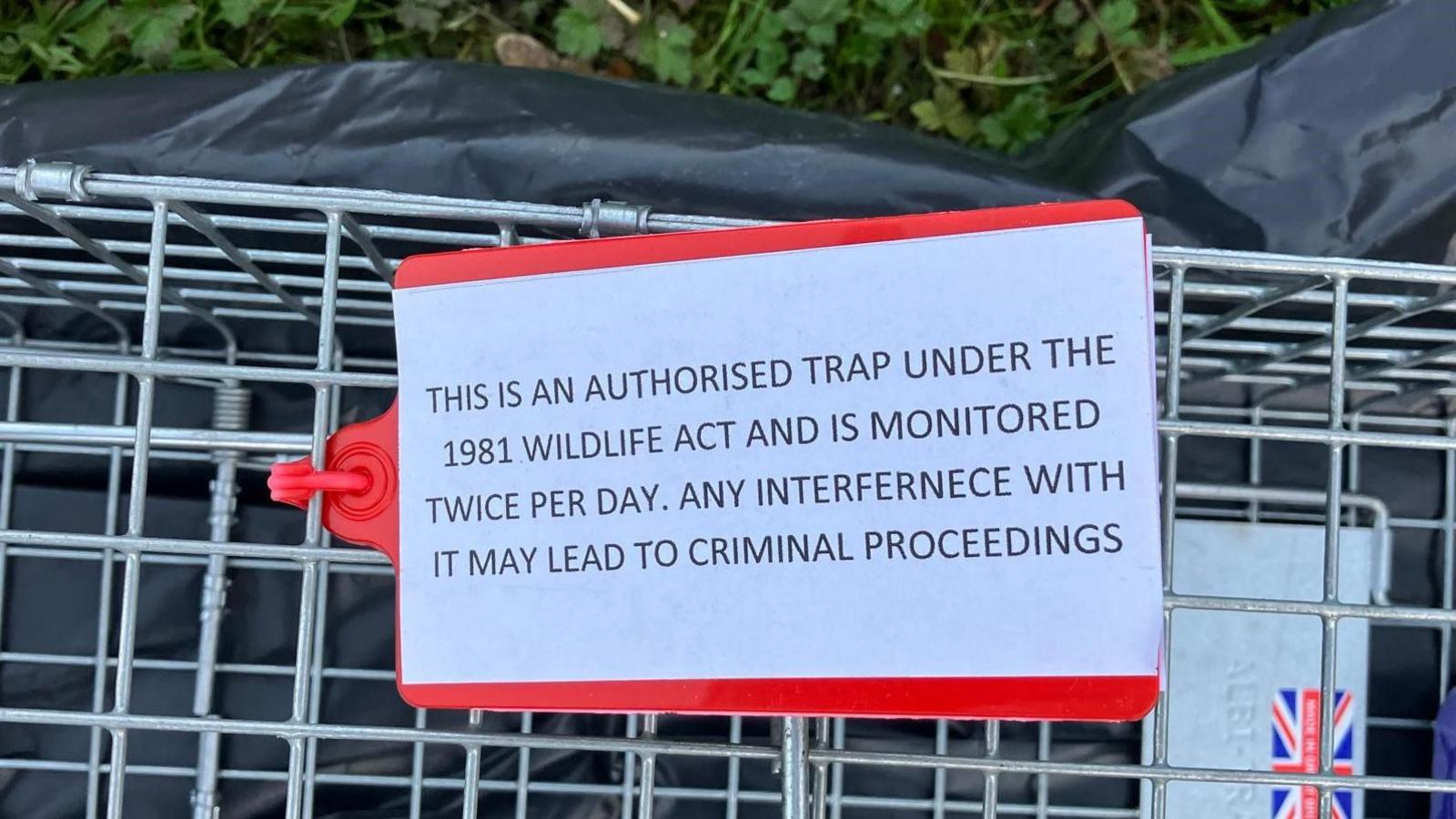 The picture shows a cage with a clear sign on it saying that the trap is authorised and monitored and interference could lead to criminal proceedings 