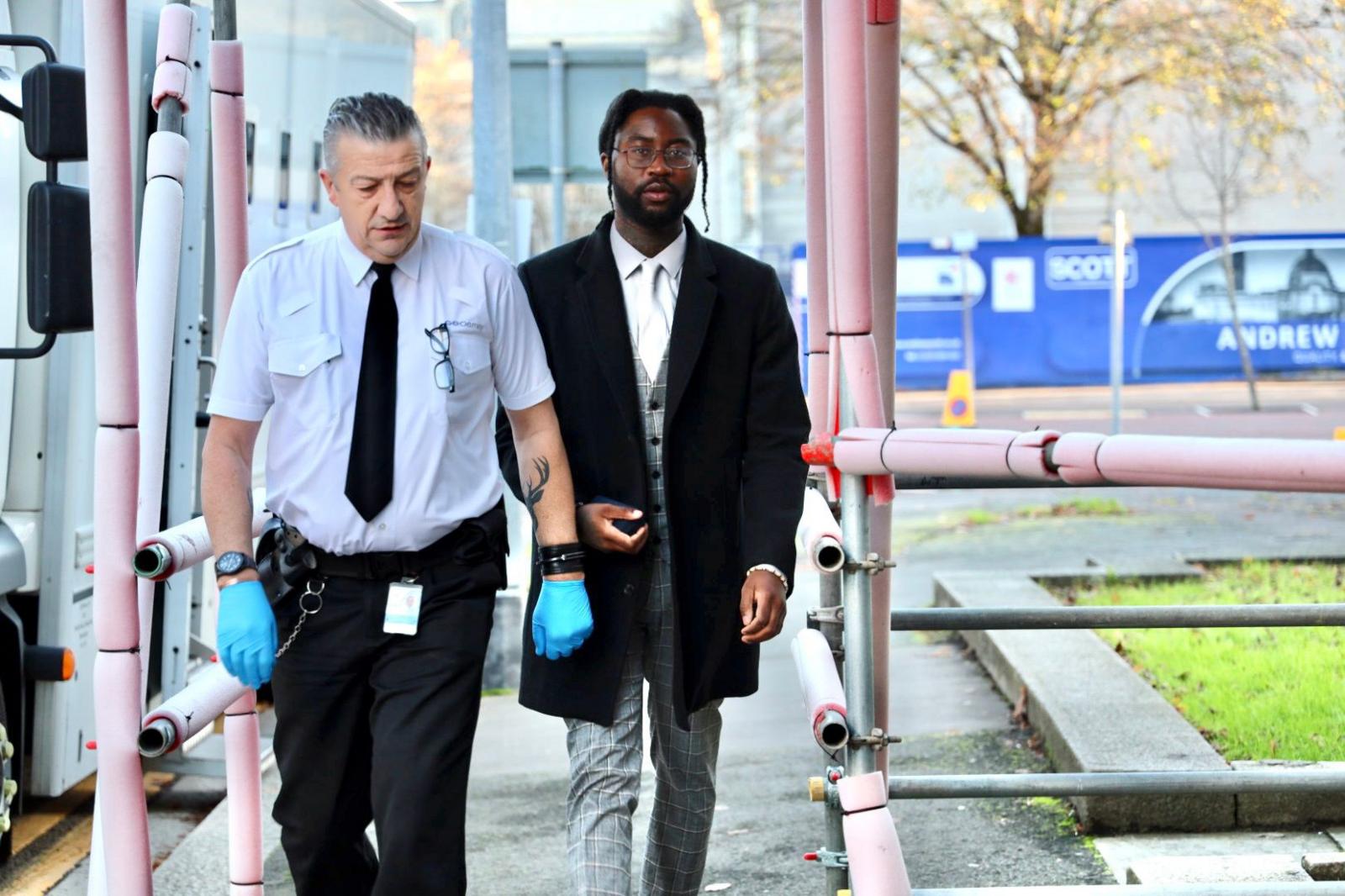 Joshua Penney arriving at court wearing glasses, a black jacket and a suit. He is accompanied by a prison warden with a white shirt and black tie. The warden wears blue gloves.
