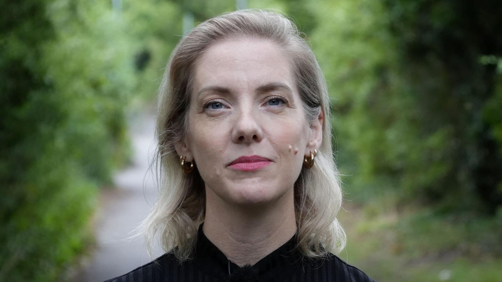 Karen Whybro has shoulder-length blonde hair and is wearing a black top while smiling. She is standing on a path flanked by bushes and trees.