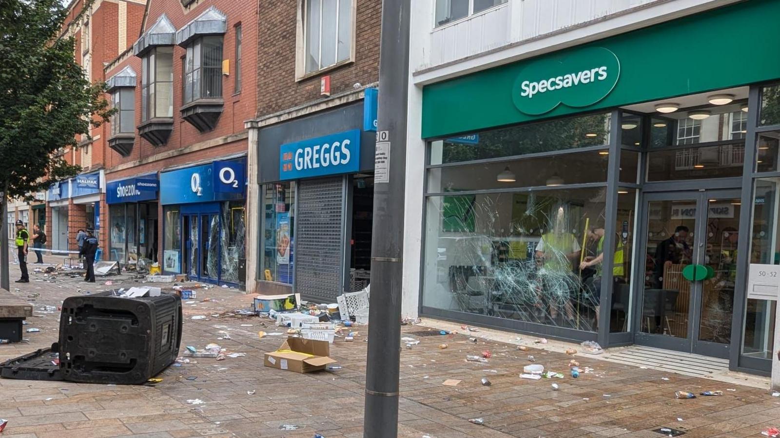 Stores with damaged glass fronts and rubbish strewn on the pavement. A black upturn wheelie bin lies on the ground in the bottom left.