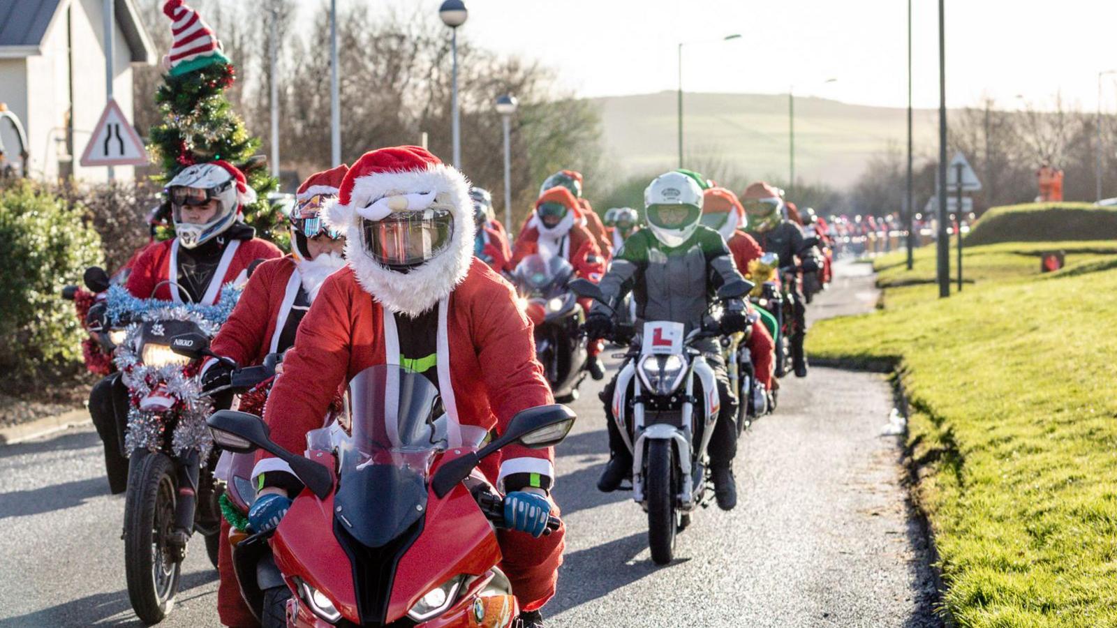 A convoy line of motorcycle riders driving towards the camera, with each rider wearing a red and white Santa Claus costume. Some motorcycles are also decorated with Christmas lights and tinsel. 
Hills are in the background. 