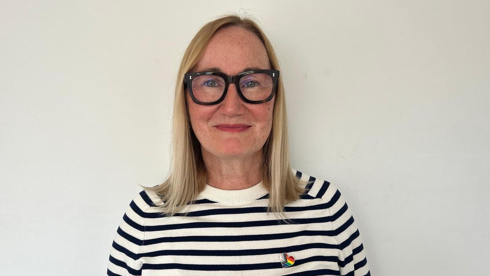 A head and shoulders shot of Anya. She is wearing a black and white striped jumper and has a Pride pin on her right lapel. She has shoulder length blonde straight hair and is wearing thick framed black glasses. 