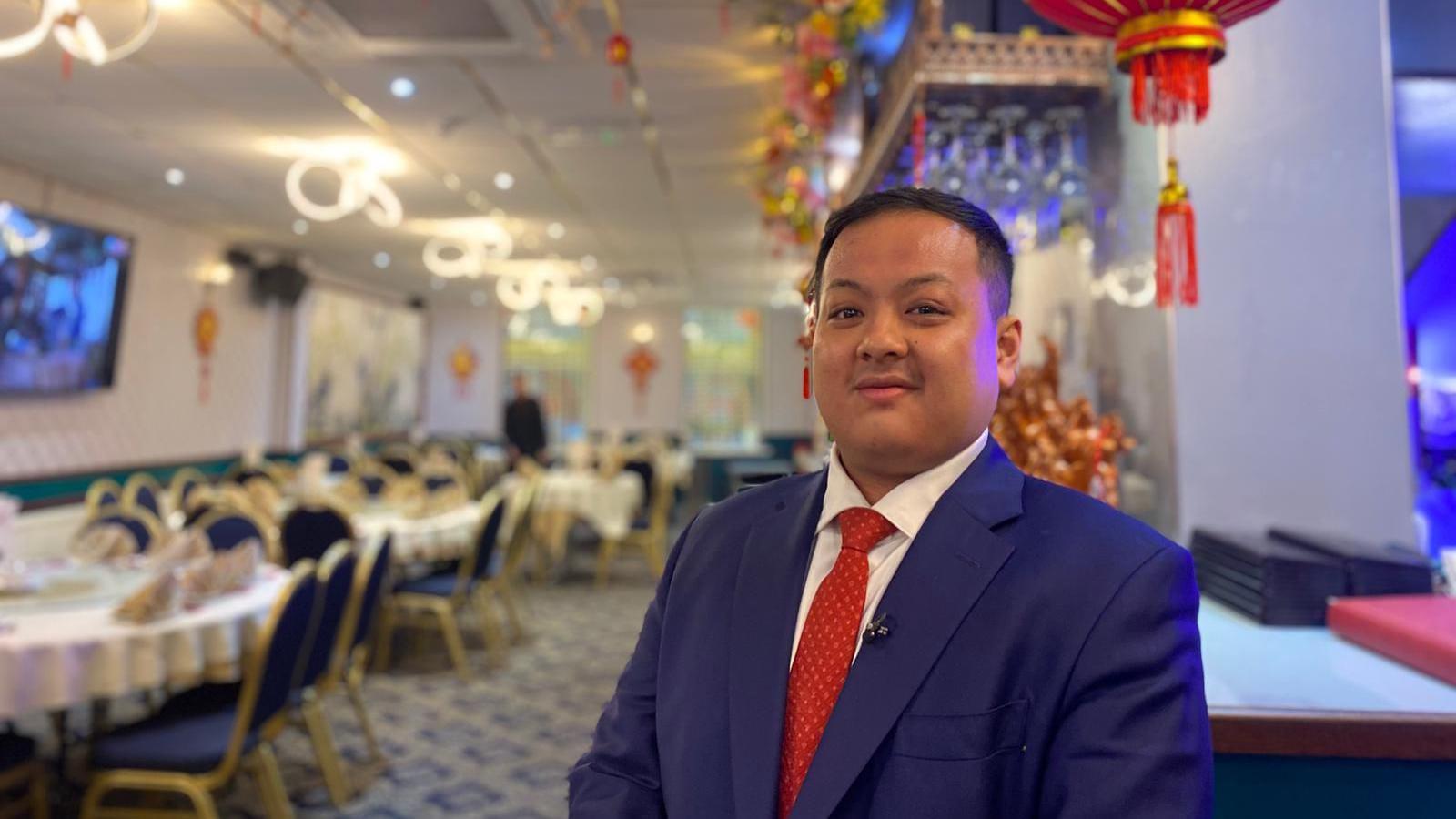 A Chinese man wears a navy blazer, red tie and white shirt, and smiles at the camera as he stands in a restaurant.