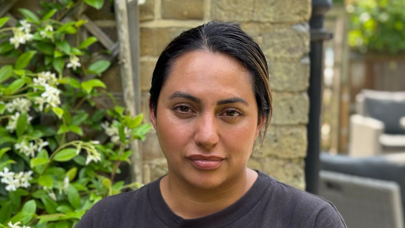 Amelia Palmer stands wearing a black T-shirt in front of brick wall and a plant with flowers to the left. To the right there is garden furniture in the distance. 