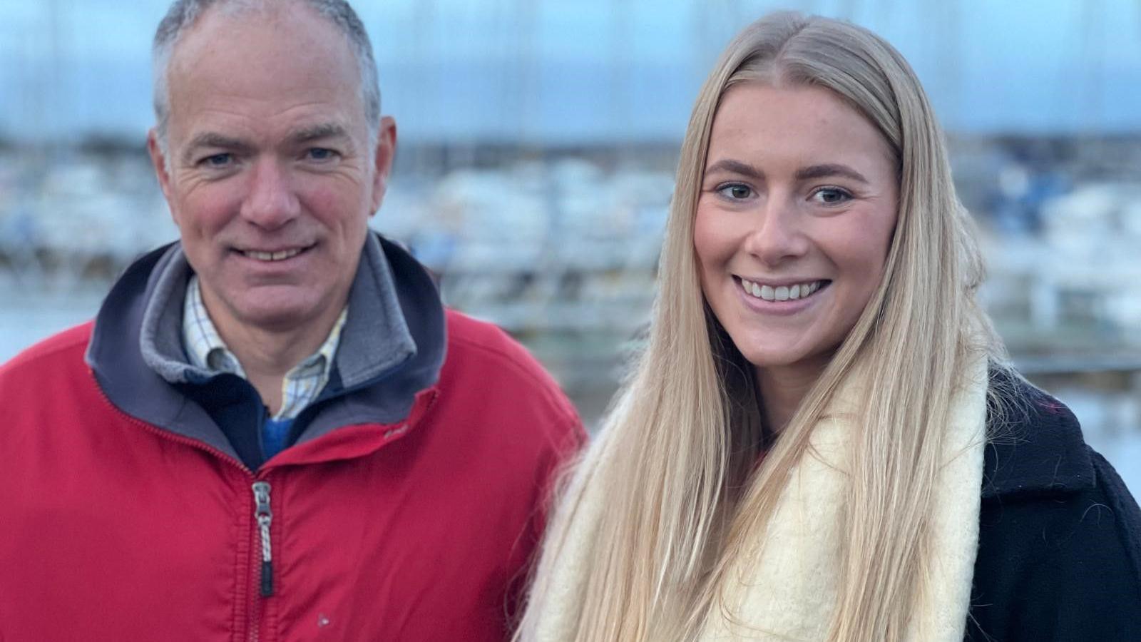 Adam Waugh and Natasha Dawson. They are posing together and smiling at the camera. Mr Waugh has grey short hair and is wearing a red jacket. Natasha Dawson has long blond hair and is wearing a white scarf and a black coat.