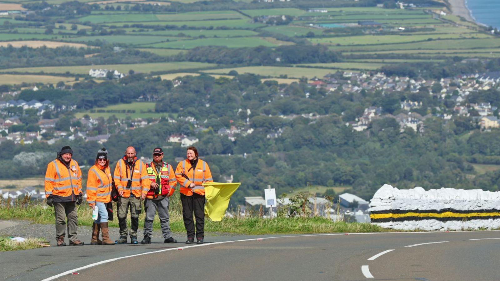 Four marshals in orange high vis jackets standing on the Mountain Road with Ramsey below in the background. One marshal is holding a yellow flag, and there is a stone wall painted white with yellow and balck stripes to the right.