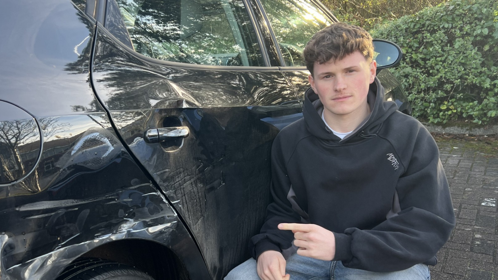 Joshua Durant crouching next to his black Seat Ibiza pointing to the damage.