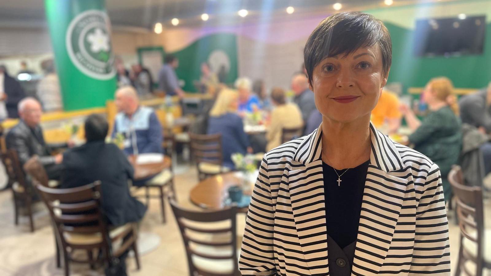 A woman with a black and white stripped blazer is smiling in a cafe. She has a black top on underneath her blazer and a grey waistcoat. She has short hair and is wearing lipstick.