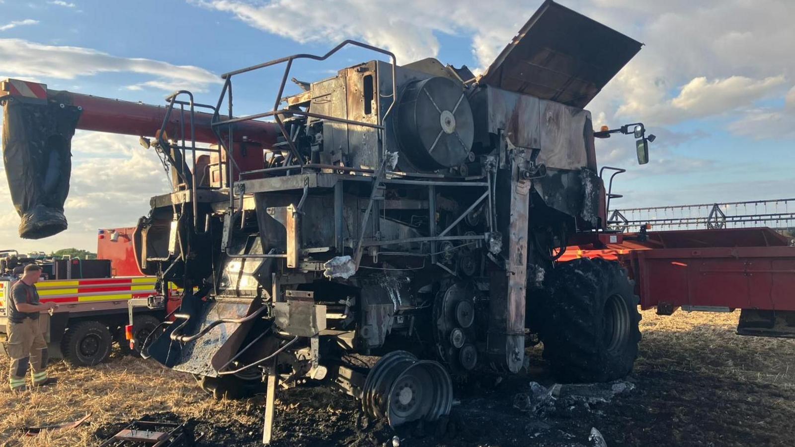 A close-up of a burnt out combine harvester. Its main body is blackened, its front wheel burnt to the rim. It is sitting in blackened field stubble. To the left is a firefighter looking at a flatbed fire service lorry, in red and yellow fire service livery, which is just beyond the combine.