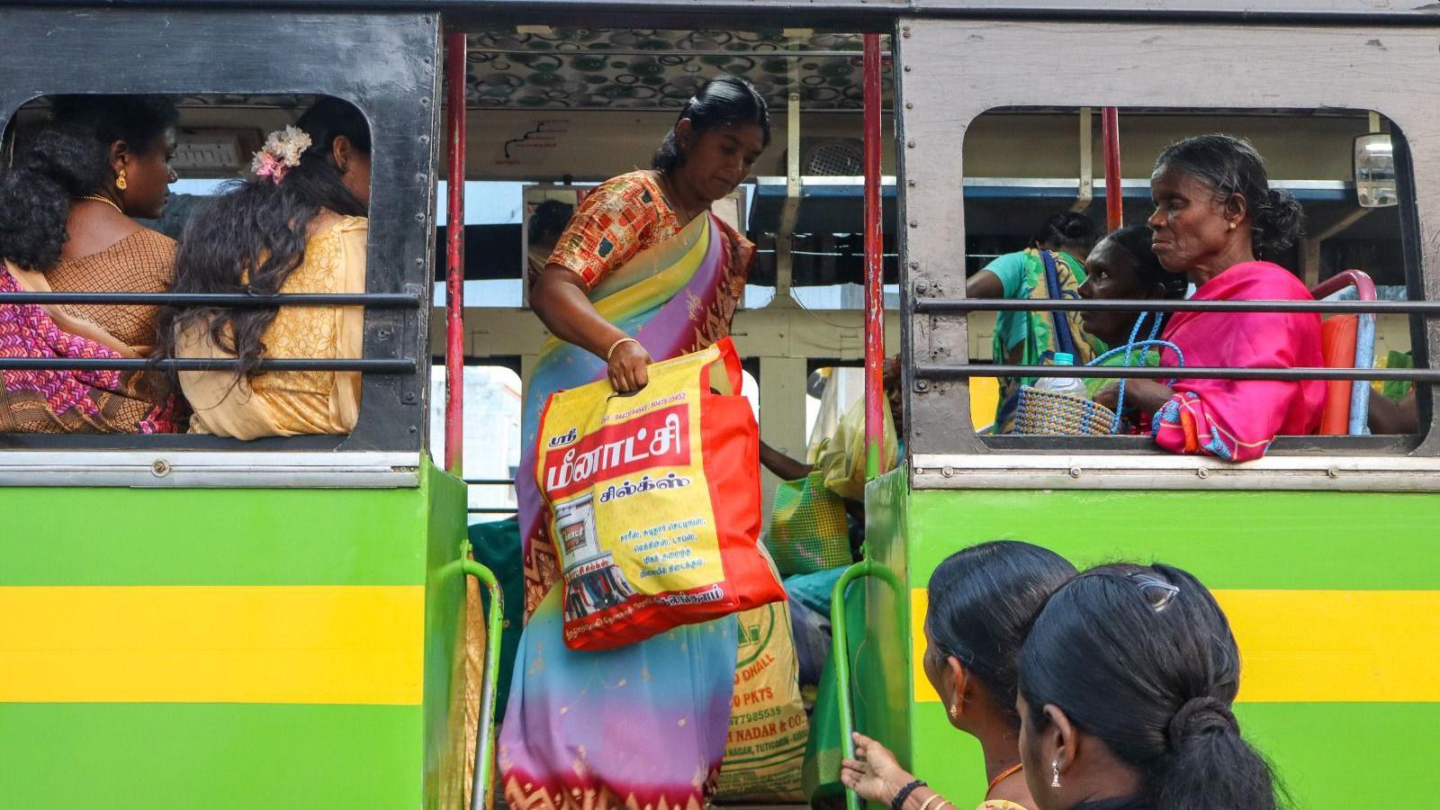 A woman wearing a multicoloured saree gets out of a green coloured public bus while holding a yellow and red bag filled with goods. 