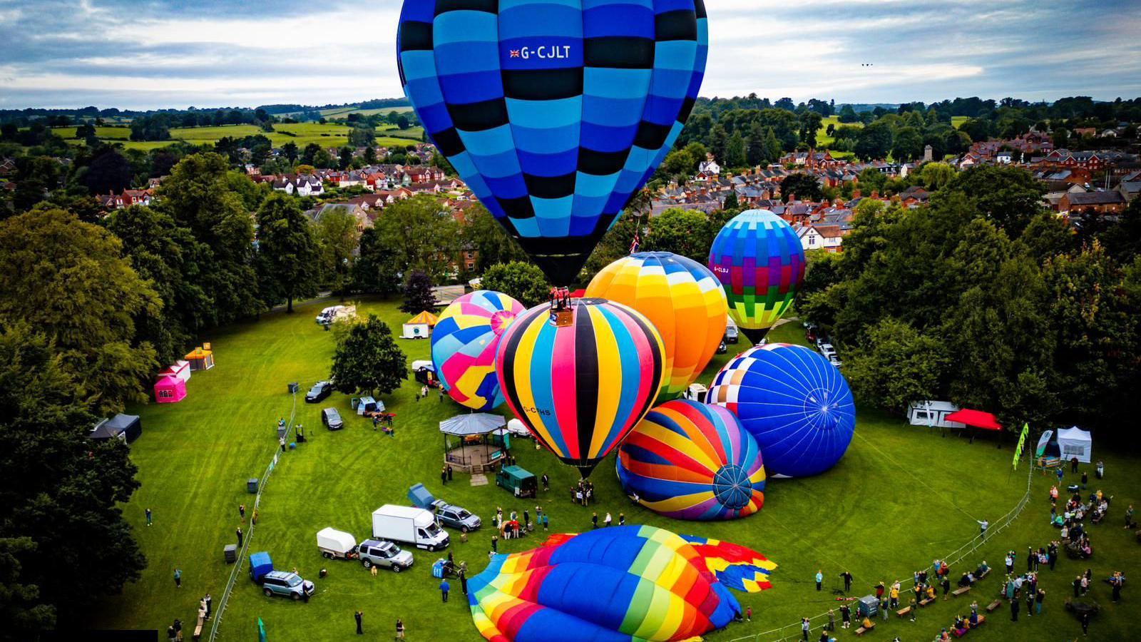 An aerial shot of the balloons taking off. Several balloons are on the ground inflating while one blue and black balloon is rising in the foreground of the shot. 