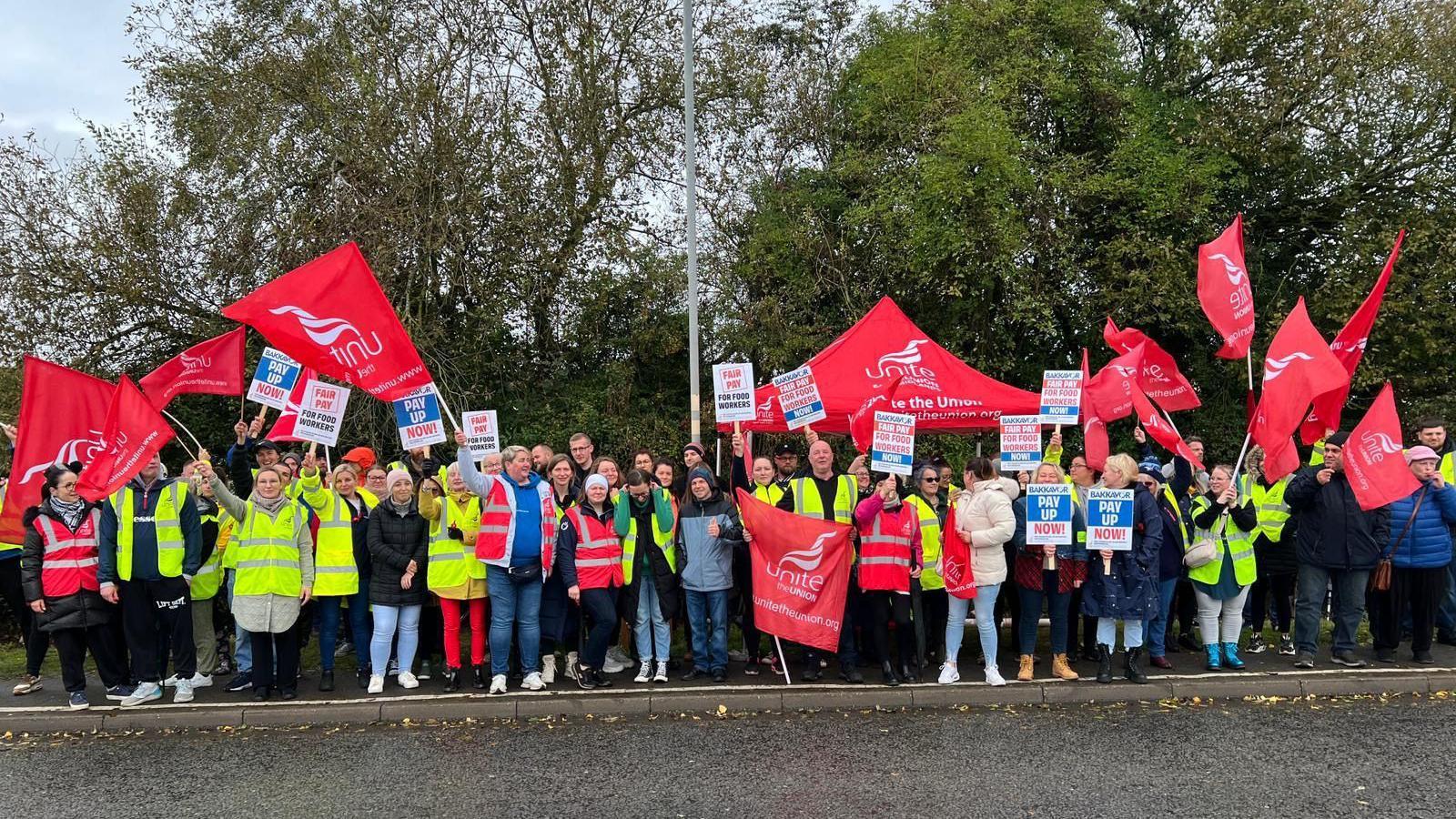 A crowd of striking workers outside the Spalding factory. They are mainly wearing hi-vis jackets and have Unite the Union flags and placards.