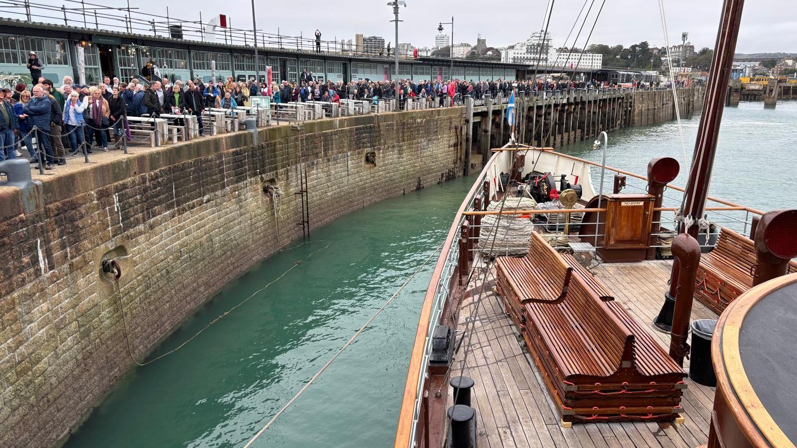 Shot taken from on board Waverley paddle steam showing the bow of the vessel and crowds lining Folkestone Harbour Arm