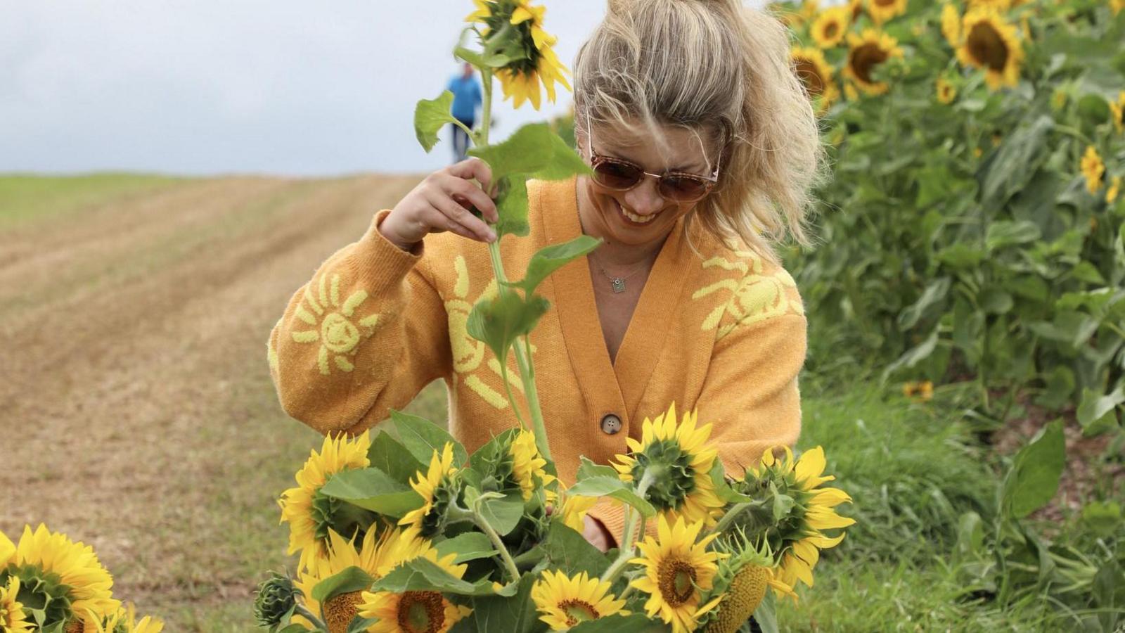 A woman wearing sunglasses and an orange cardigan with stylised images of the sun on it is arranging a bouqet of sunflowers. There is a field of sunflowers behind her.