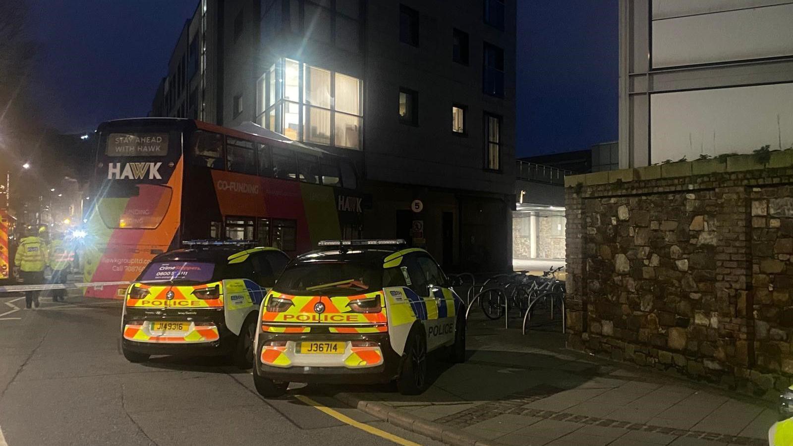 A lime green, black and pink double-decker bus crashed into a building overhang, with a crumpled top deck and debris lying in the road while it is still dark. There are two police cars parked nearby and police officers can be seen near to the bus.