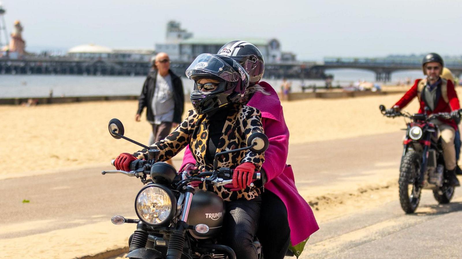 A woman in leopard print jacket riding a black motorbike with a woman in a pink  jacket on the back as a passenger.