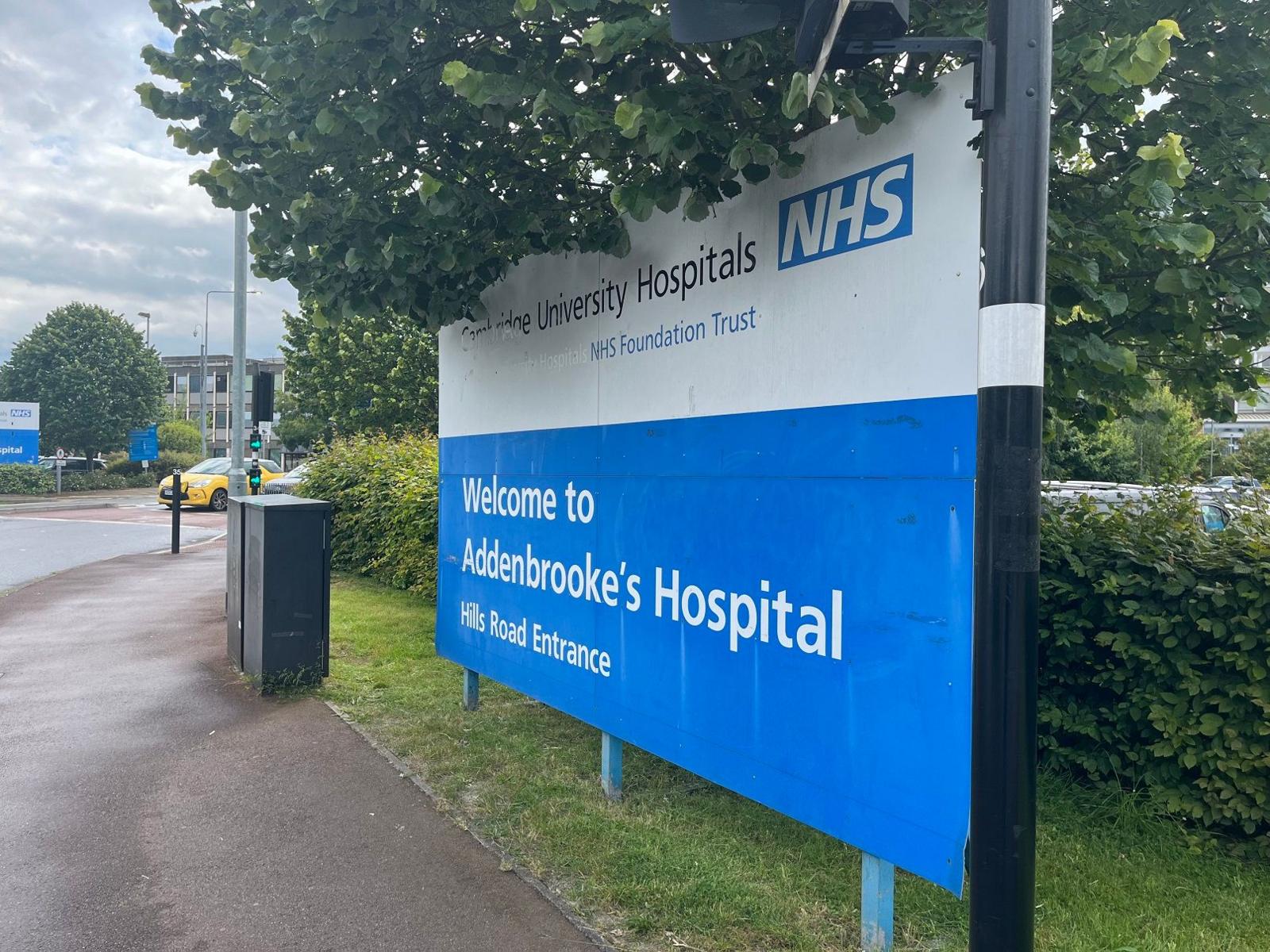 The blue and white Welcome sign at Addenbrooke's Hospital, next to a hedge and under a tree. In the distance is a car park and the hospital building.
