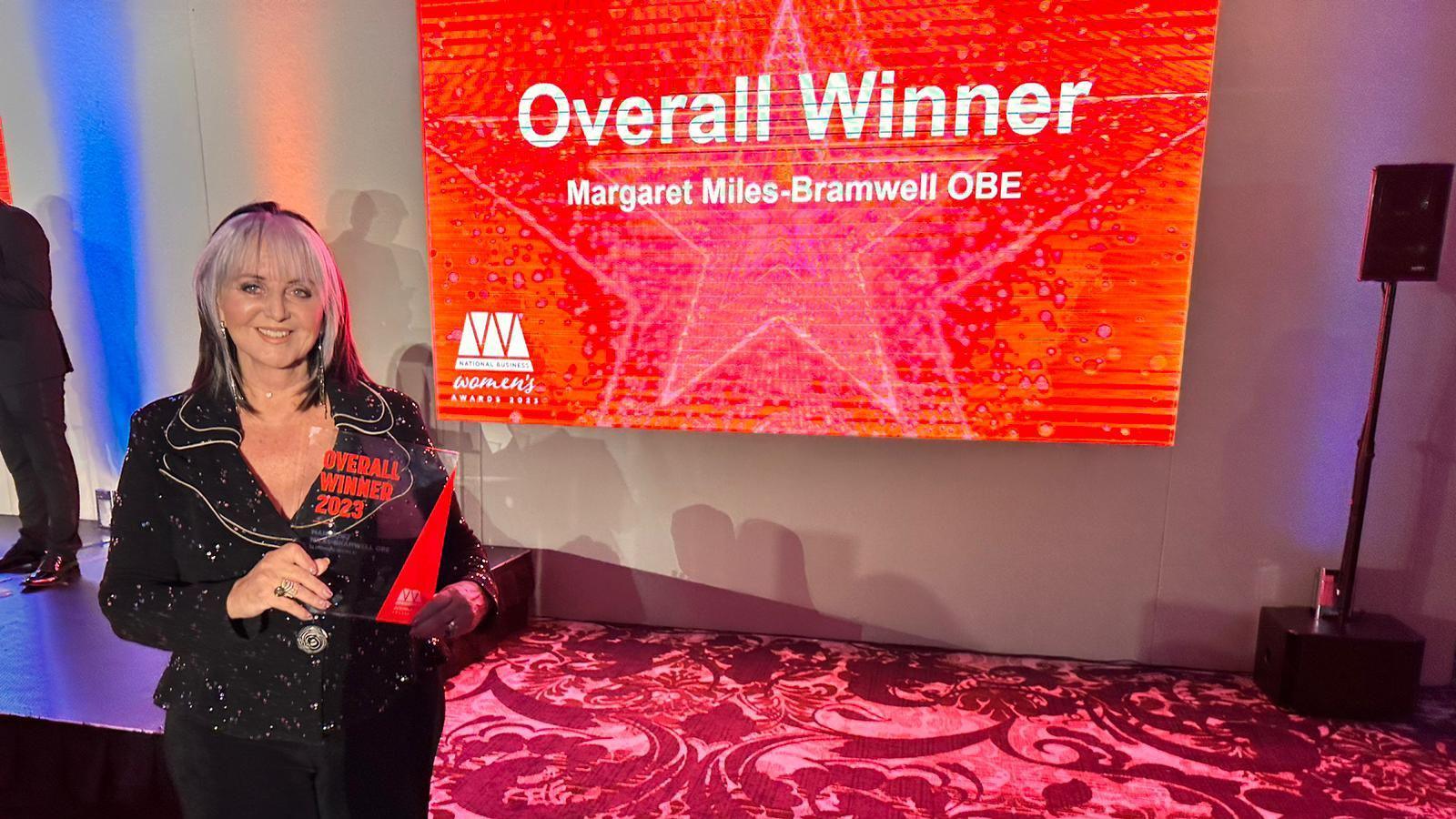 Mrs Miles-Bramwell holding a trophy, and behind her is a screen saying "overall winner Margaret Miles-Bramwell". 