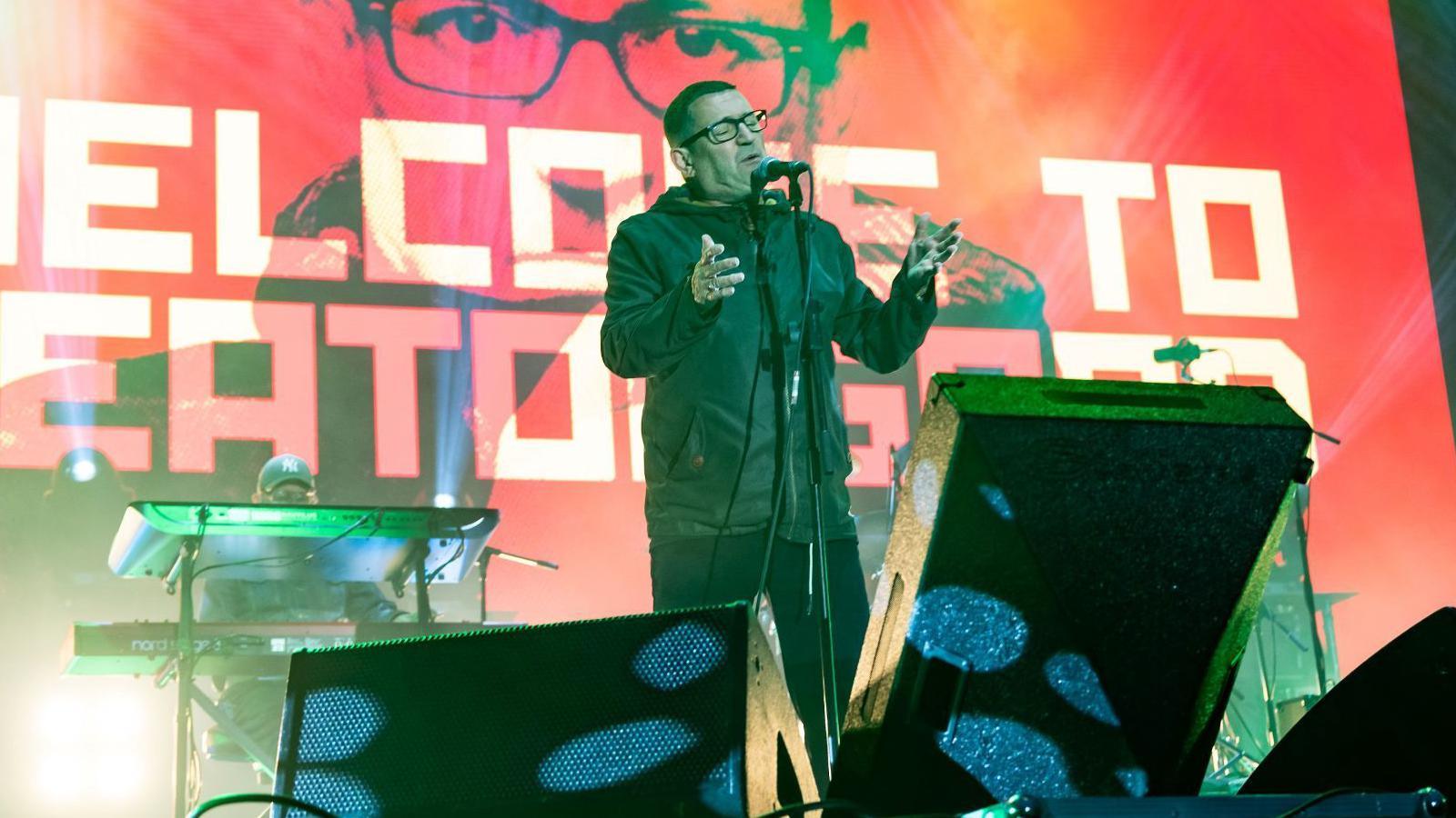 Paul Heaton has a shaven head and is wearing glasses and a green hooded jacket. He has his eyes closed as he sings into a microphone.