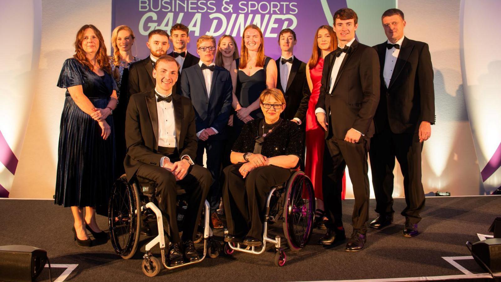 A group of young people standing together, two wheelchair users, smiling at the camera. All are wearing black tie attire and are in front of a large banner saying Wiltshire Business & Sports Gala Dinner.