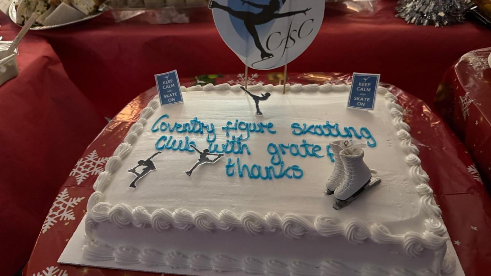 A white rectangle cake with icing decoration around the outside and miniature skating figures and ice skates. Writing from blue icing in the centre of the cake reads 'Coventry figure skating club with grateful thanks'.