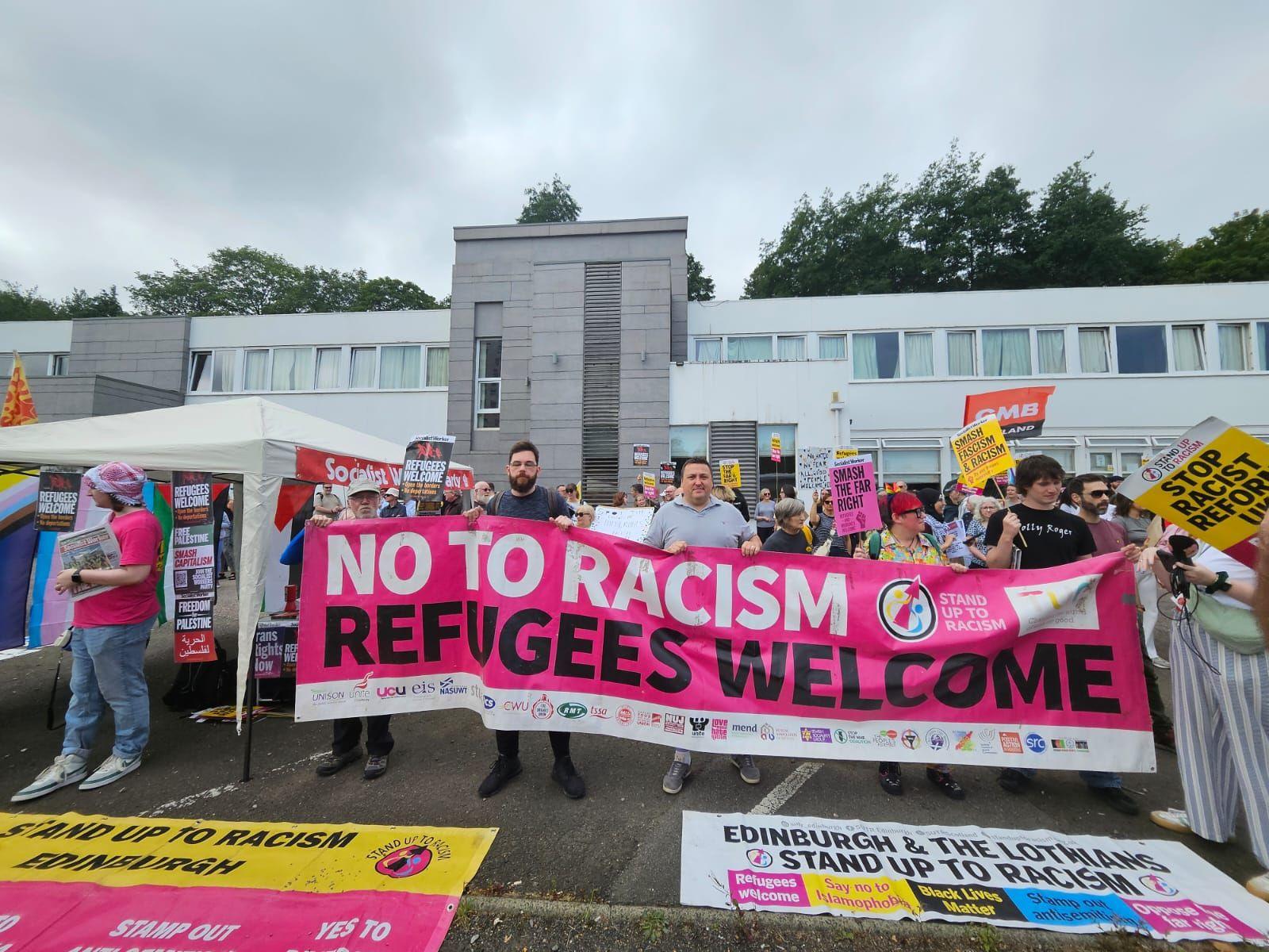 Protesters holding a large banner saying No to Racism, Refugees Welcome outside a building