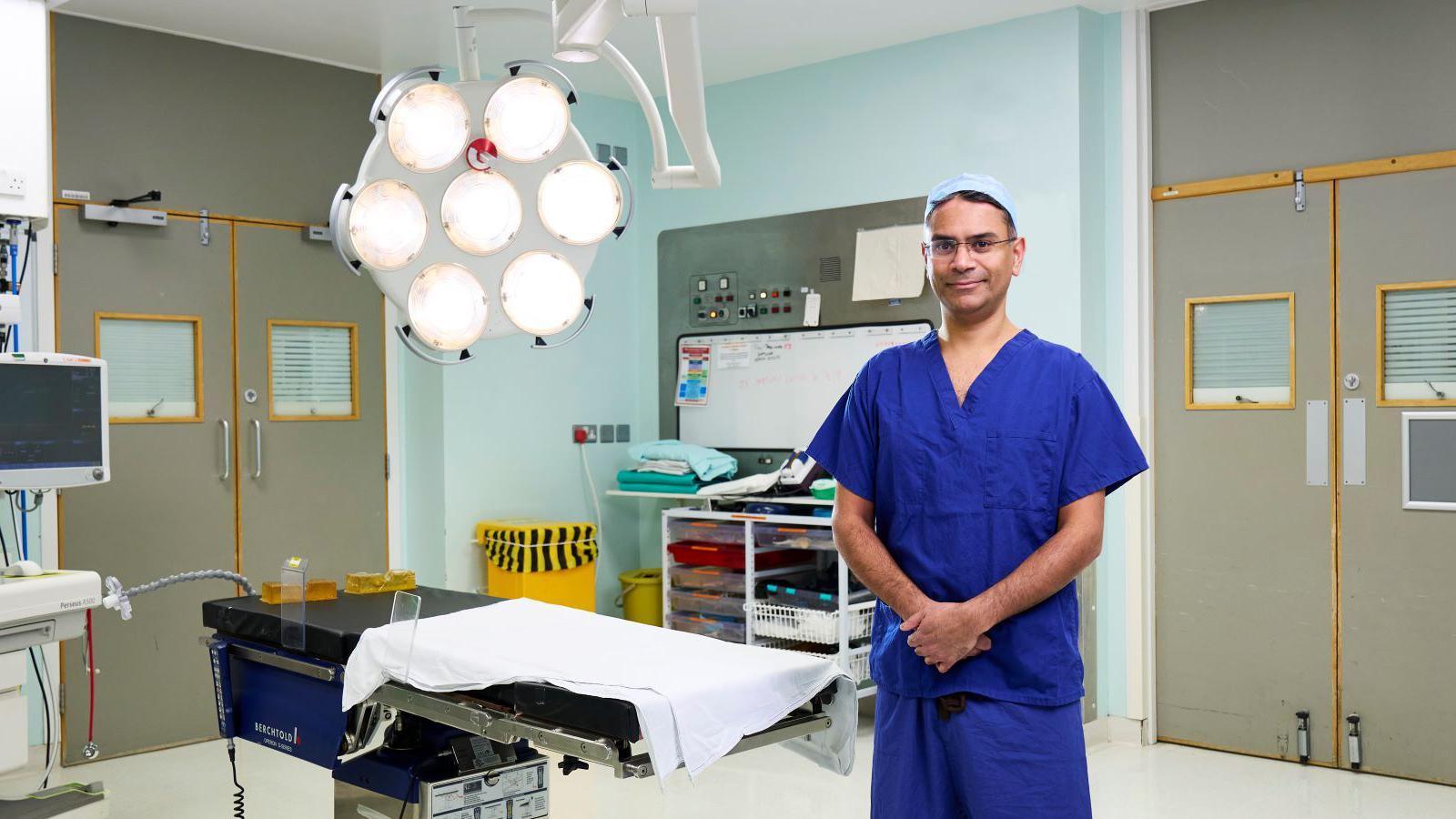 Mr Patel is standing in an operating theatre wearing blue scrubs. His hands are clasped in front of him. he is wearing glasses and is smiling at the camera.