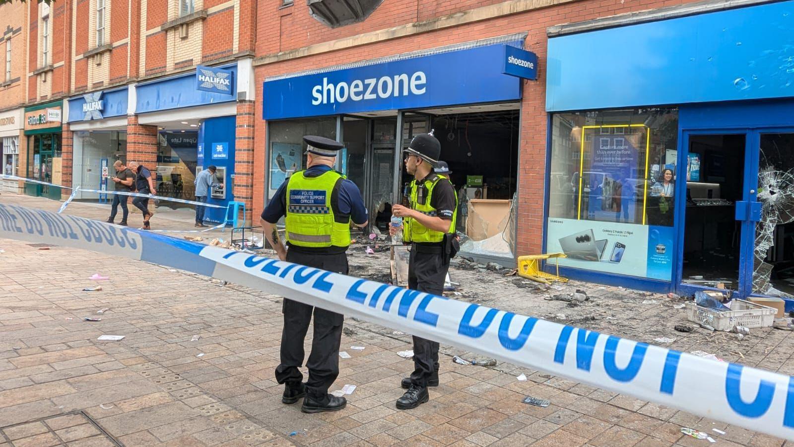 Two police officers standing behind a cordon and in front of damaged shops with debris scattered on the ground