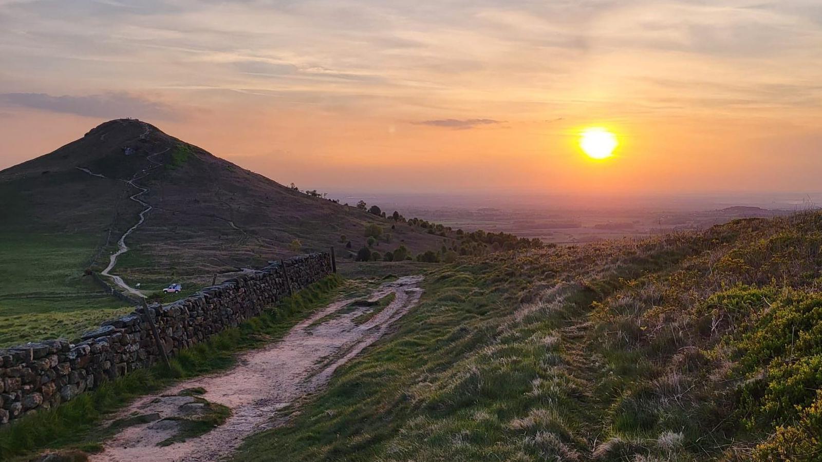 A sunset from the top of Roseberry Topping. A stone wall runs alongside a path running up the peak of the hill in the distance.