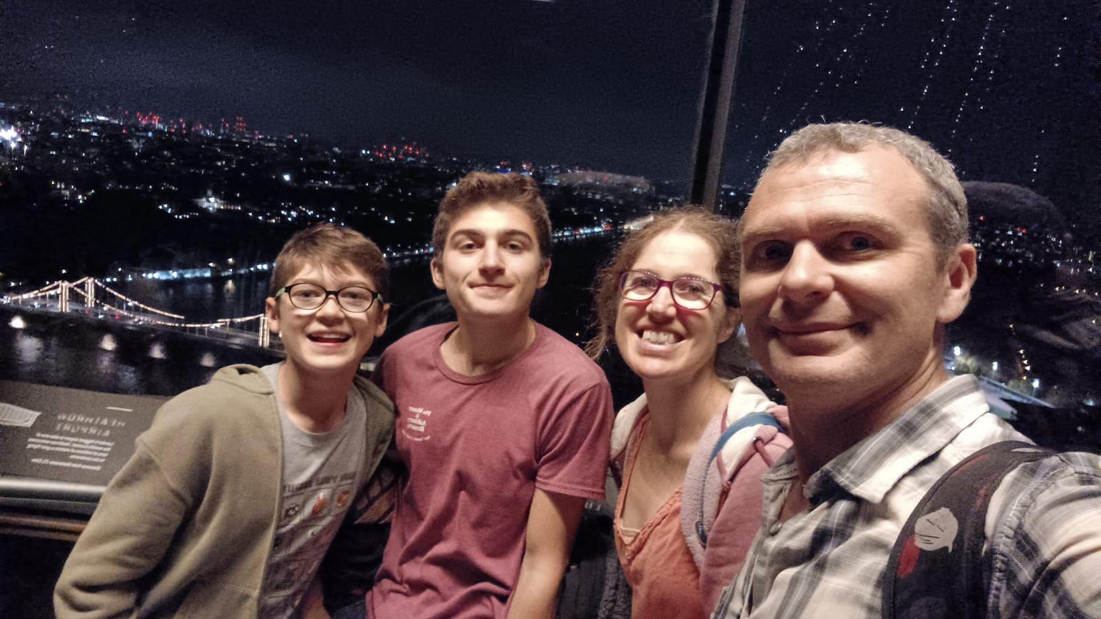 A selfie of a man with grey wearing a blue and white checked shirt. He stands beside a woman with glasses and coral t-shirt, and two young boys with brown hair. One is wearing a red t-shirt and another is wearing glasses with a grey t-shirt and green jacket.