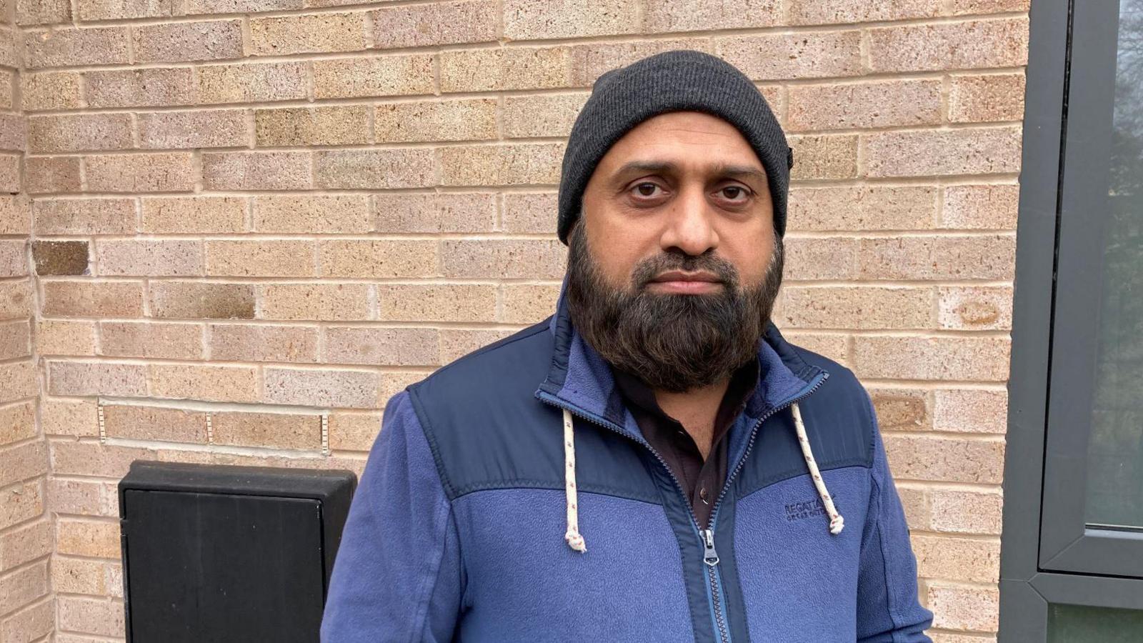 A man stands outside with a brick wall in the background. He has a black beard and is wearing a black woollen hat and a blue hooded top.