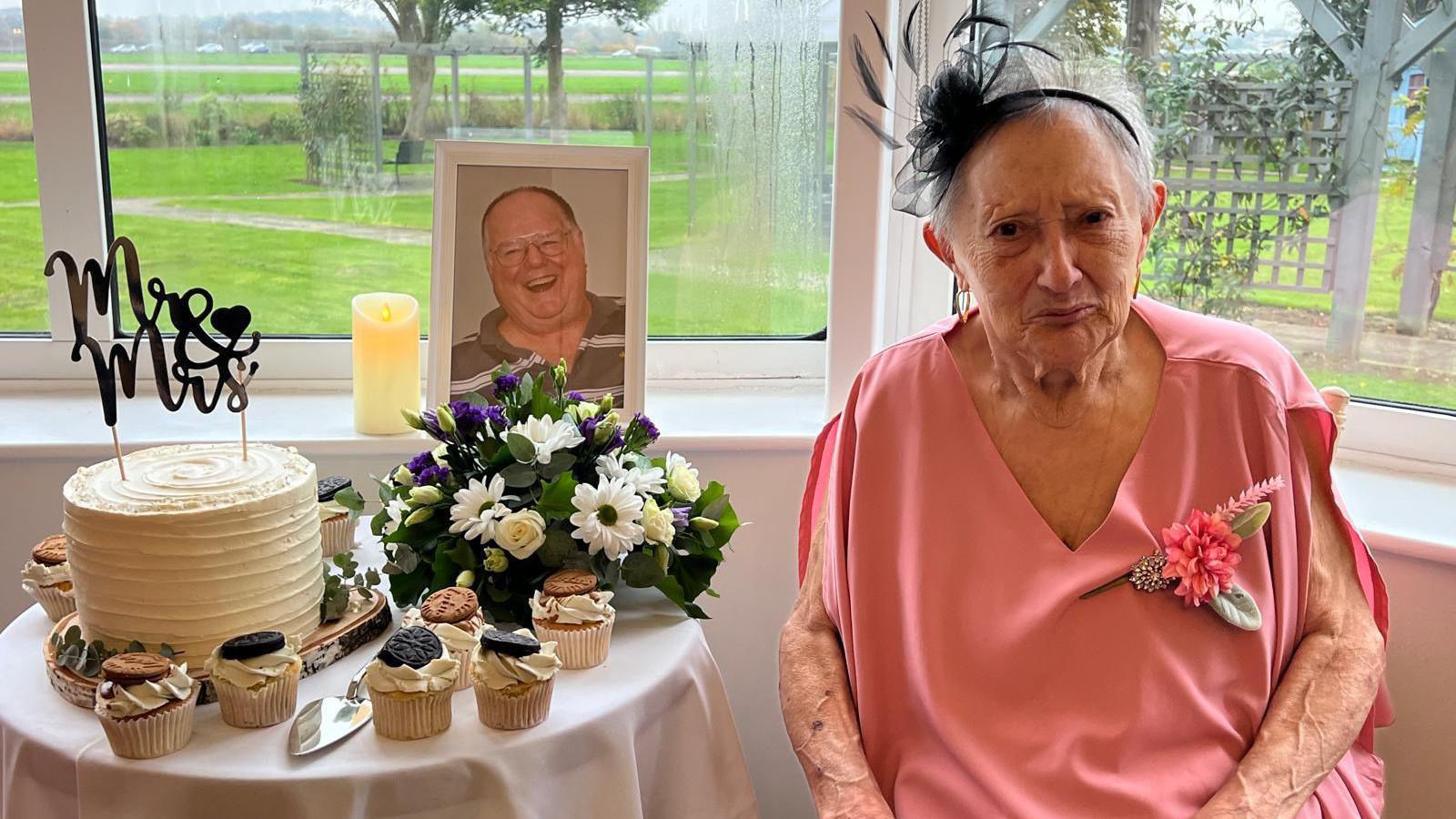 Mrs Harris is pictured sitting on a chair next to a small table with a wedding cake and several cupcakes. A picture of her husband Ken sits on the windowsill behind her. She is smiling at the camera.